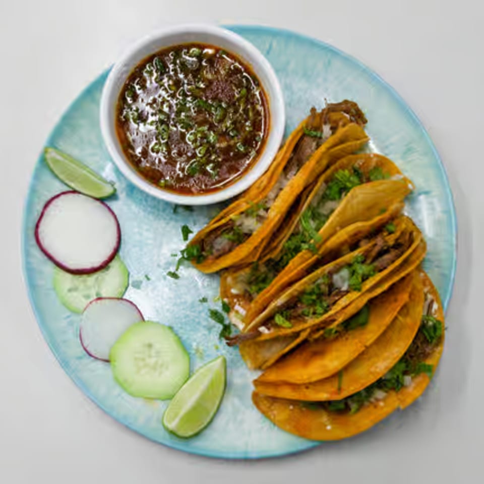 Toortugas Mexican Torteria's birria tacos with a side of consommé, sliced radishes, cucumbers, and lime.