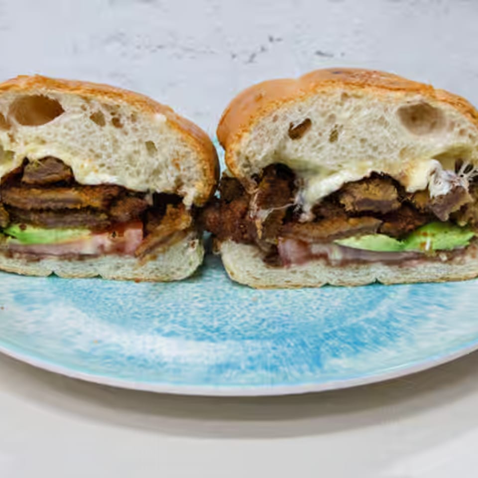 A Toortugas Mexican Torteria sandwich, cut in half, revealing layers of meat, avocado, and cheese.