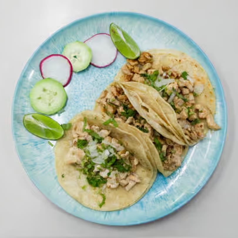 Three chicken tacos with cilantro and onions, served with lime, radish, and cucumber slices.