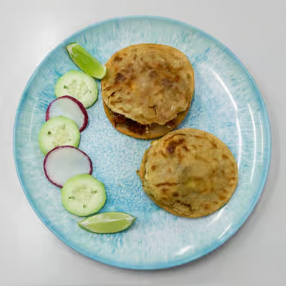 Two Mexican gorditas with a side of sliced cucumbers, radishes, and lime wedges on a blue plate.