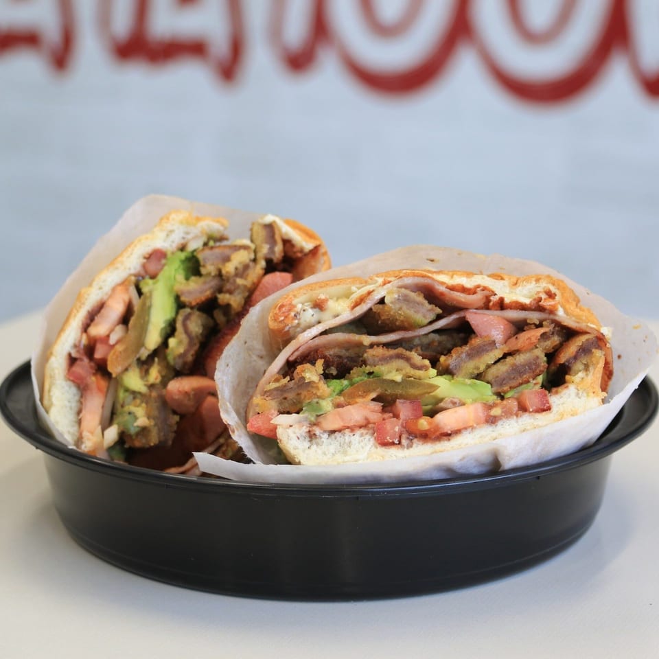 Two Mexican tortas, filled with meat, avocado, tomatoes, and hot dogs, in a black bowl.