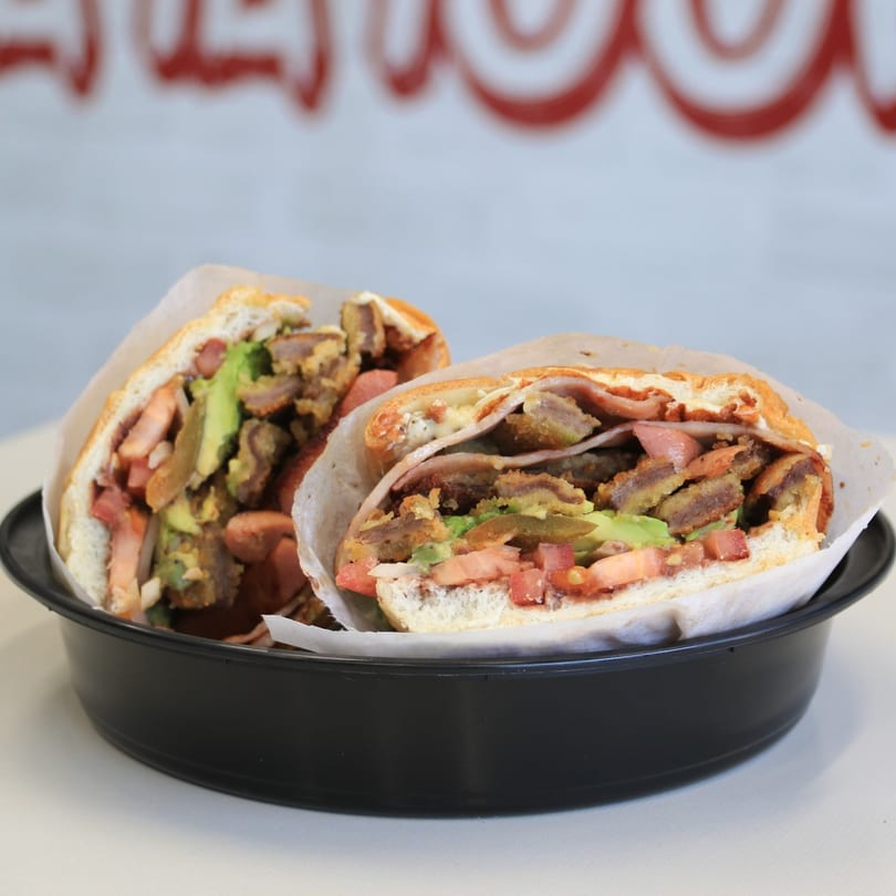 Two Mexican tortas, filled with meat, avocado, tomatoes, and hot dogs, in a black bowl.