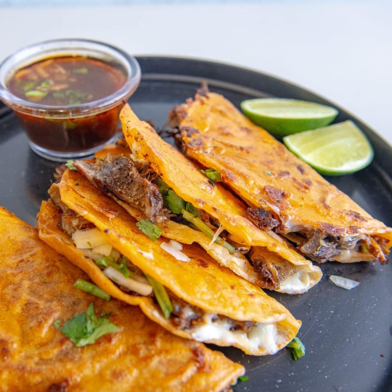 Toortugas Mexican Torteria birria tacos with consommé and lime wedges on a black plate.