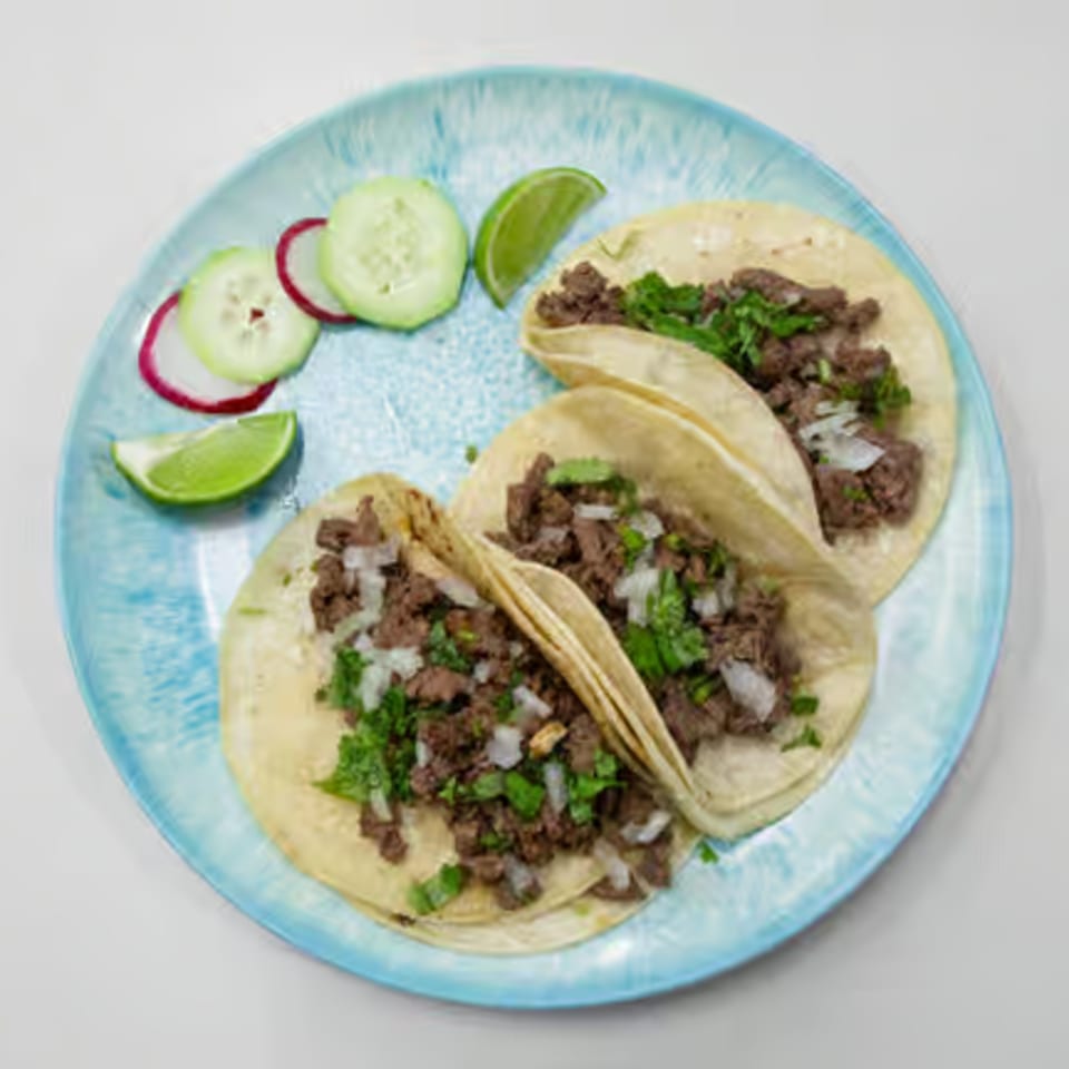 Three Toortugas Mexican Torteria tacos with meat, onions, cilantro, and lime on a blue plate.